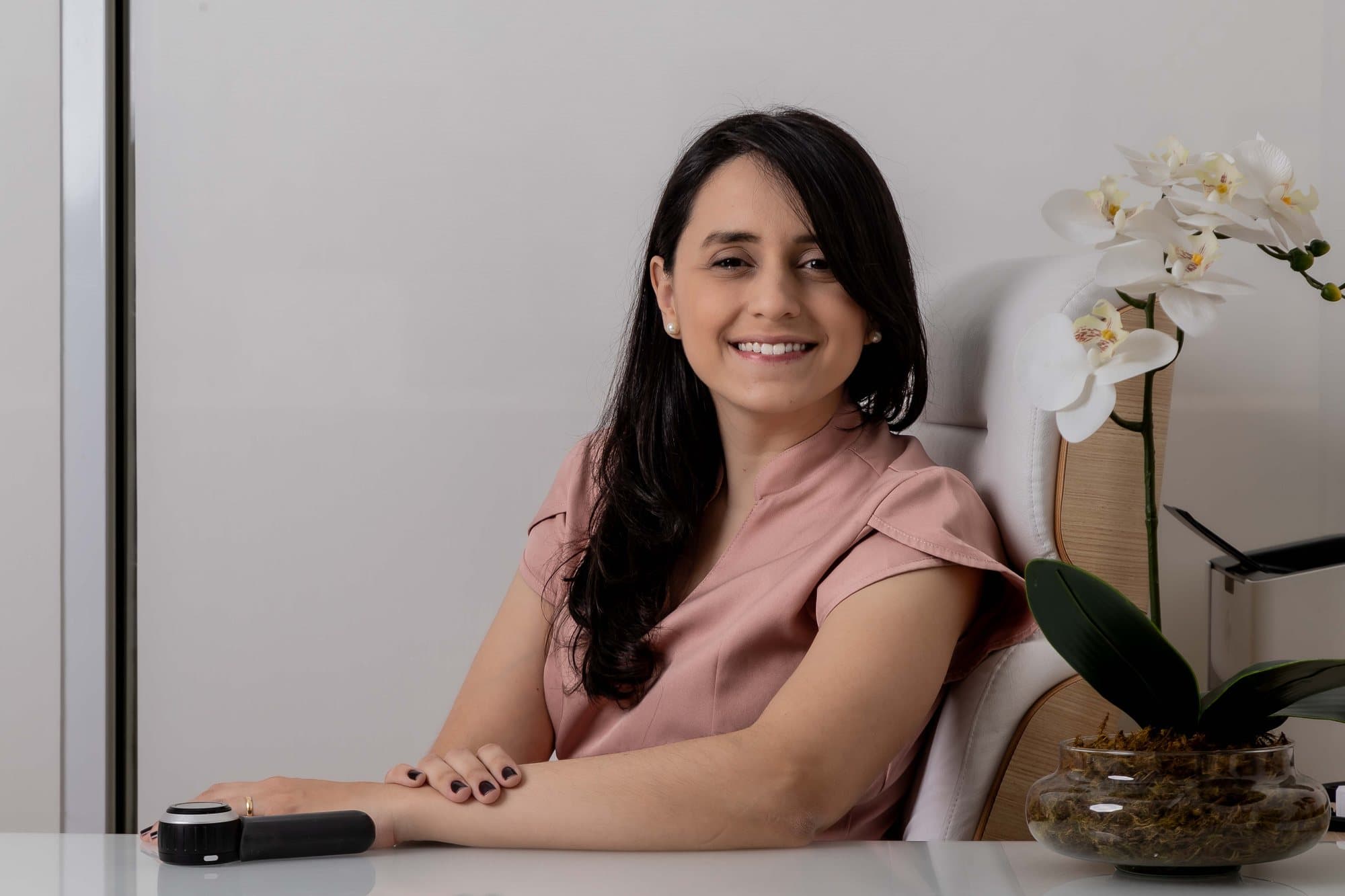 Dra. Paula Azevedo sorrindo à mesa do consultório, com o dermatoscópio ao lado